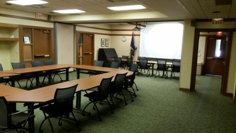 Picture of a meeting room with opened dividers down the middle and a projector screen on the far wall. There are two sets of tables surrounded by chairs, one near and one farther next to the projector screen.