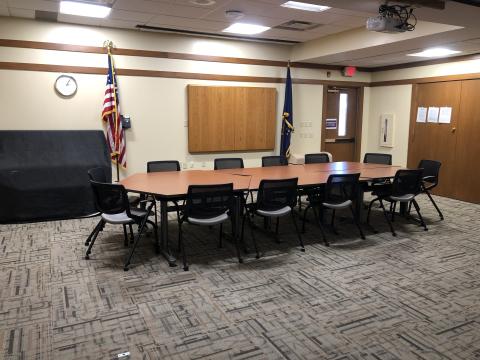 Picture of a meeting space with several small tables arranged to form a long single table surrounded by chairs. There is a projector mounted in the ceiling and a door on the back wall.