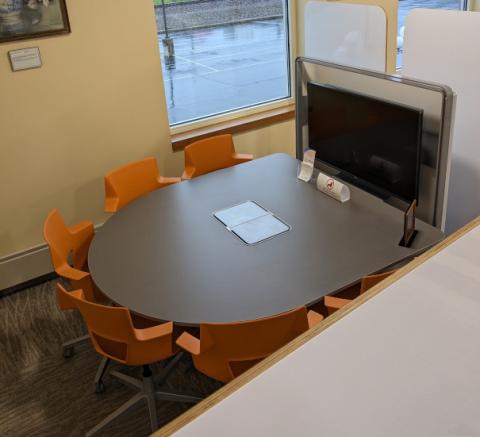 Image of a half circle table surrounded by chairs on the round side and a television on the flat side with privacy panels all around.